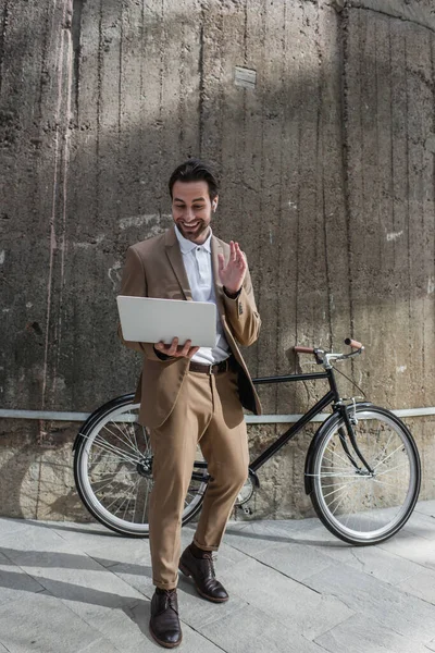 Pleine longueur d'homme d'affaires heureux dans les écouteurs tenant ordinateur portable tout en ayant appel vidéo à l'extérieur — Photo de stock