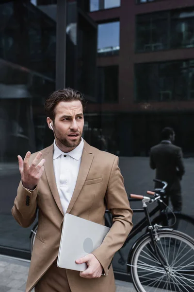 Homme d'affaires dans les écouteurs parlant et tenant un ordinateur portable près du bâtiment et du vélo — Photo de stock