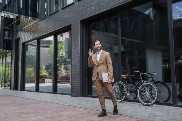 Pleine longueur d'homme d'affaires heureux dans les écouteurs agitant la main et tenant un ordinateur portable près du bâtiment et du vélo — Photo de stock