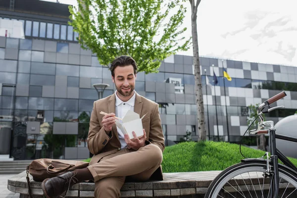 Happy businessman in suit holding cardboard box with asian food and chopsticks near bike — Stock Photo