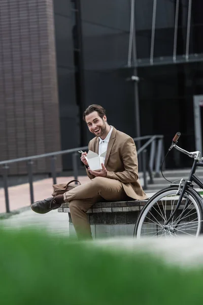 Empresário feliz em terno segurando caixa de papelão com comida asiática e pauzinhos perto de bicicleta — Fotografia de Stock