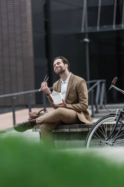 Задоволений бізнесмен у костюмі тримає картонну коробку з азіатською їжею та паличками біля велосипеда на вулиці — стокове фото
