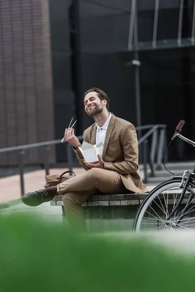 Heureux homme d'affaires en costume tenant boîte en carton avec de la nourriture asiatique et baguettes près de vélo à l'extérieur — Photo de stock