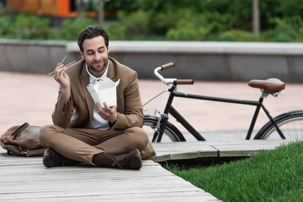 Happy businessman in suit holding cardboard box with asian food and chopsticks while sitting with crossed legs — Stock Photo