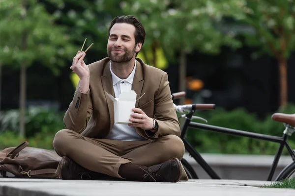 Pleased businessman in suit holding chopsticks and asian food while sitting with crossed legs — Stock Photo