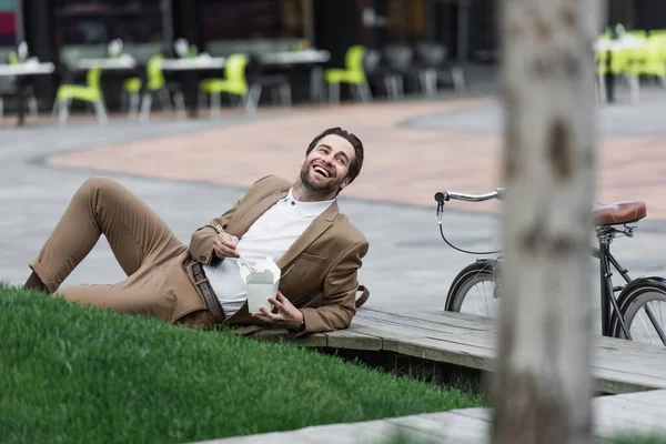 Happy businessman in suit holding cardboard box with asian food and chopsticks while lying near grass — Stock Photo