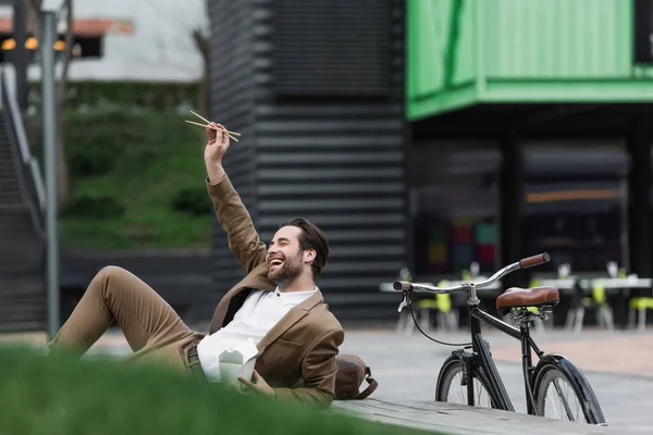 Alegre hombre de negocios en traje sosteniendo caja de cartón con comida asiática y palillos mientras está acostado cerca de la hierba - foto de stock