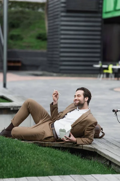 Homem de negócios feliz segurando caixa de papelão com comida asiática e pauzinhos enquanto deitado perto de grama — Fotografia de Stock