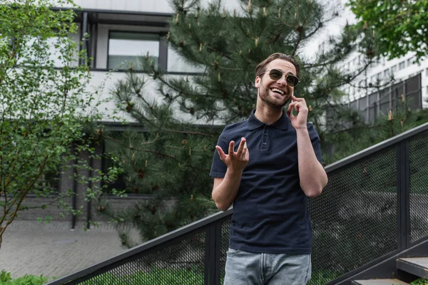 Fröhlicher Mann mit Sonnenbrille und Polohemd, der draußen mit dem Handy telefoniert — Stockfoto