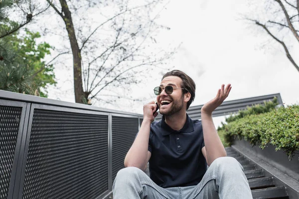 Uomo sorridente con occhiali da sole e polo che parla al cellulare fuori — Foto stock