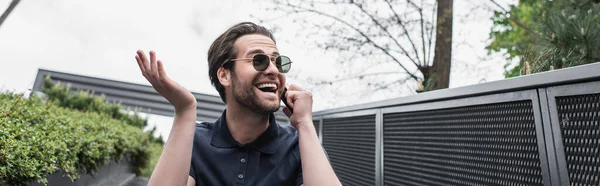 Hombre sorprendido en gafas de sol y polo hablando en el teléfono celular exterior, pancarta - foto de stock