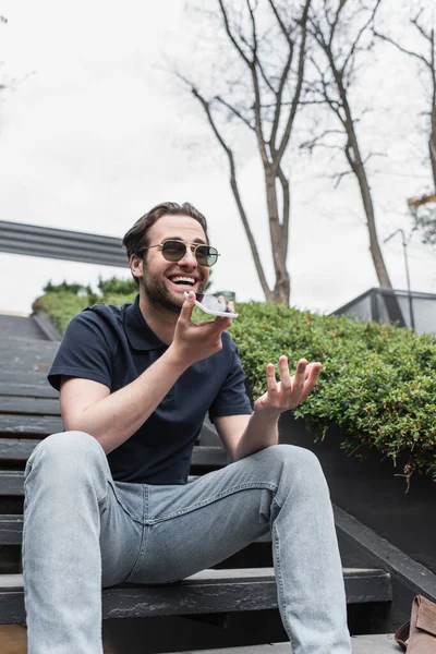 Cheerful man in sunglasses and polo shirt recording voice message on cellphone outside — Stock Photo