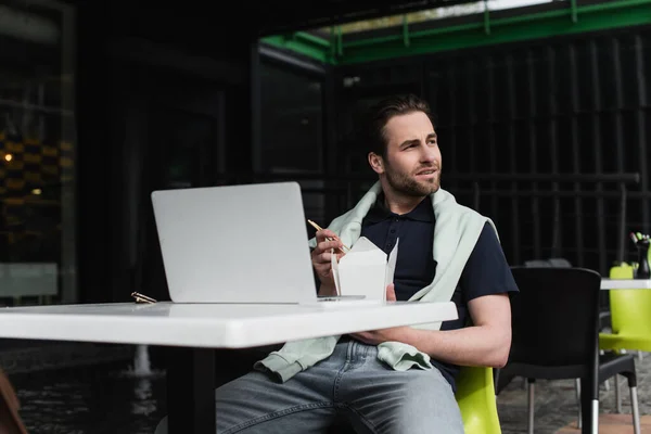 Bearded man in polo shirt and sweatshirt holding chopsticks and carboard box while sitting in outdoor cafe — Stock Photo