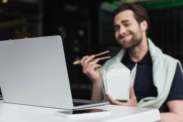 Hombre desenfocado en polo y sudadera sosteniendo palillos y caja de cartón mientras mira el portátil cerca del teléfono inteligente - foto de stock