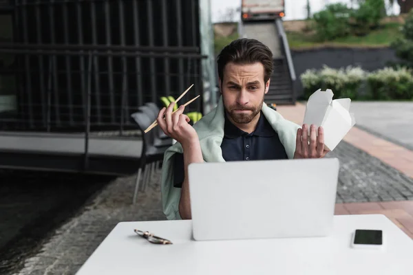 Homem barbudo em camisa de pólo e camisola segurando pauzinhos e caixa de papelão enquanto olha para laptop perto de smartphone na mesa — Fotografia de Stock
