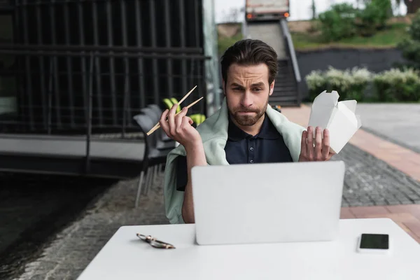 Bärtiger Mann in Poloshirt und Sweatshirt mit Essstäbchen und Kartonschachtel beim Blick auf Laptop neben Smartphone auf Tisch — Stockfoto