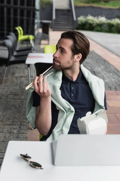 Bearded man in polo shirt and sweatshirt holding chopsticks and carboard box near laptop and blurred sunglasses on table — Stock Photo