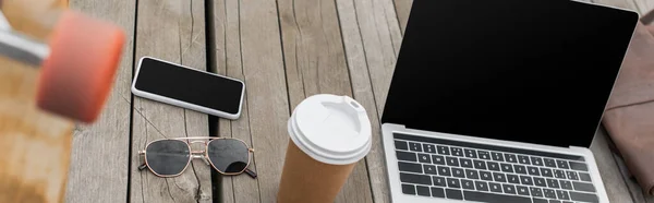 Gadgets avec écran blanc, lunettes de soleil et tasse en papier sur table en bois près de longboard flou, bannière — Photo de stock