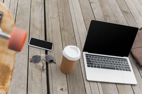 Gadgets with blank screen, sunglasses and paper cup on wooden table near blurred longboard — Stock Photo