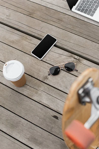 Vista ad alto angolo di gadget con schermo bianco, occhiali da sole e tazza di carta su tavolo in legno vicino al longboard sfocato — Foto stock