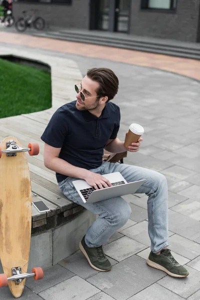 Man in sunglasses and polo shirt holding paper cup and using laptop near smartphone and longboard — Stock Photo