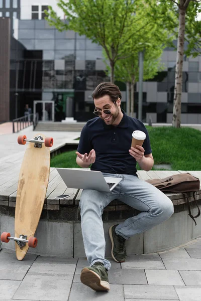 Hombre feliz en gafas de sol y polo camisa sosteniendo taza de papel y tener video chat en el ordenador portátil cerca de smartphone y longboard - foto de stock