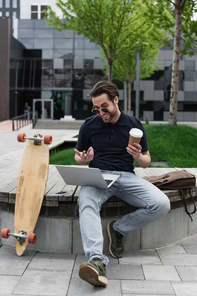 Felice uomo in occhiali da sole e polo in possesso di tazza di carta e avendo video chat su laptop vicino smartphone e longboard — Foto stock