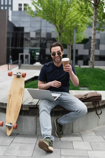 Homem feliz em óculos de sol segurando copo de papel perto de gadgets e longboard — Fotografia de Stock