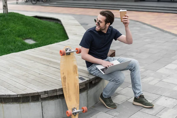 Hombre sorprendido en gafas de sol sosteniendo taza de papel cerca de gadgets y longboard fuera - foto de stock