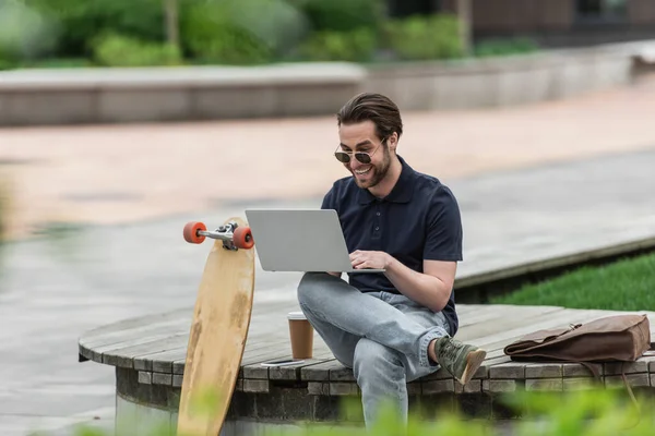 Hombre feliz en gafas de sol y polo utilizando el ordenador portátil cerca de teléfono inteligente, taza de papel y longboard - foto de stock