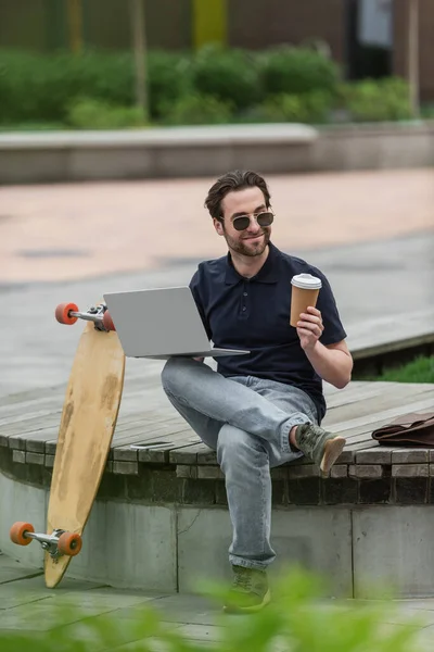 Happy man in sunglasses holding laptop and paper cup near longboard and backpack — Stock Photo