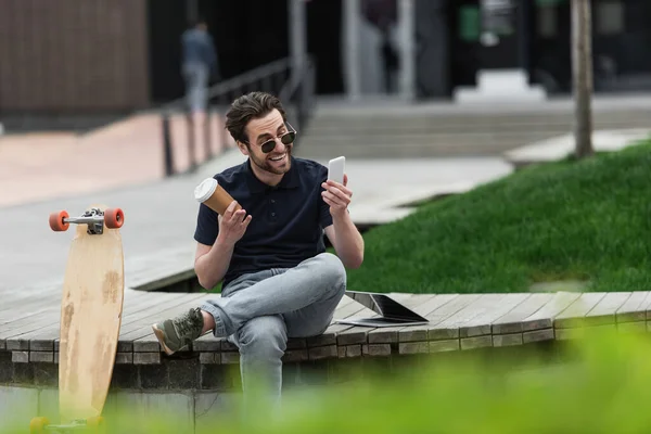 Happy man in sunglasses holding smartphone and paper cup near longboard and laptop — Stock Photo