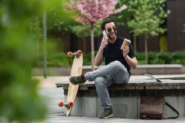 Homme heureux dans des lunettes de soleil tenant tasse en papier et parler sur smartphone près de longboard et ordinateur portable — Photo de stock