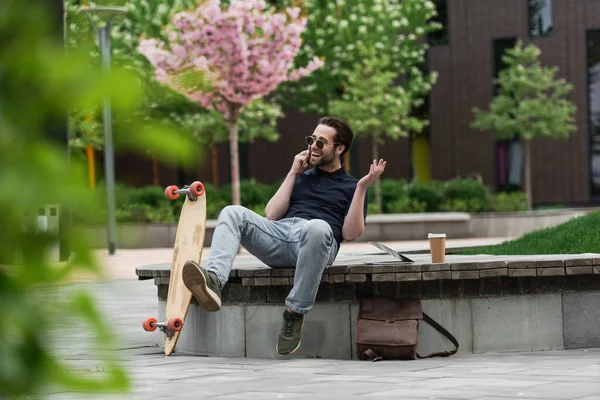 Happy man in sunglasses talking on smartphone near longboard and gadgets — Stock Photo