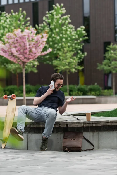 Hombre alegre en gafas de sol hablando en el teléfono inteligente cerca de longboard y gadgets - foto de stock