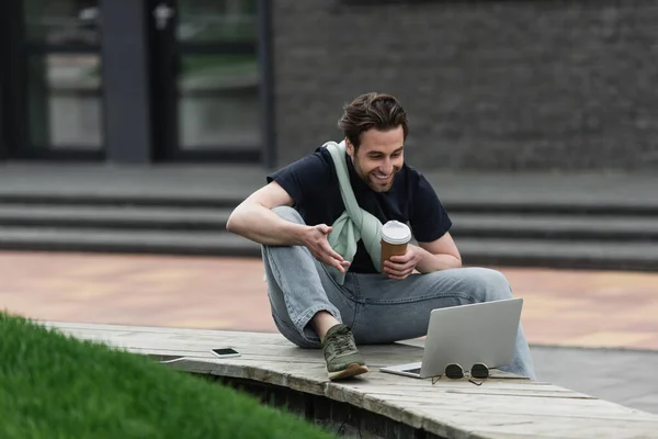 Homme gai en polo et sweat-shirt regardant ordinateur portable tout en tenant du café pour aller à l'extérieur — Photo de stock