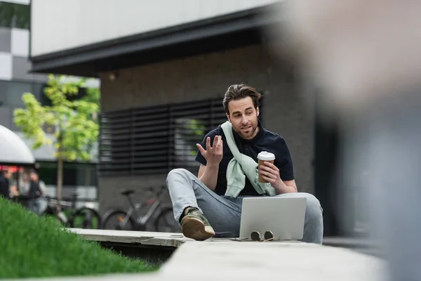 Uomo barbuto gesticolare durante la videochiamata sul computer portatile mentre tenendo tazza di carta — Foto stock