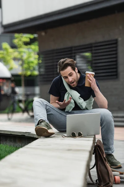 Homme barbu gestuelle pendant le chat vidéo sur ordinateur portable tout en tenant tasse de papier à l'extérieur — Photo de stock