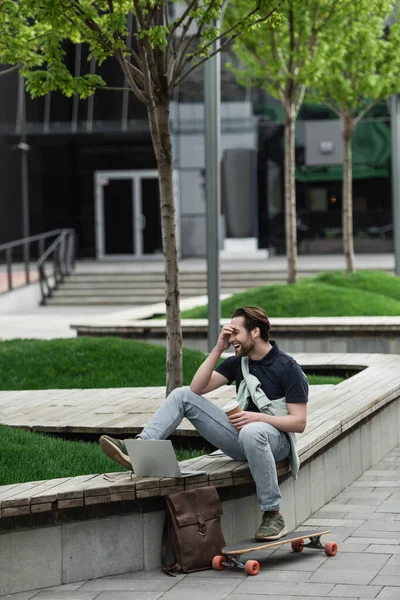 Homme heureux en polo et sweat-shirt tenant tasse en papier et riant tout en étant assis près de longboard et gadgets — Photo de stock