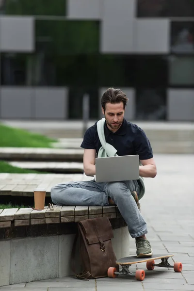 Bearded man in polo shirt and sweatshirt using laptop near paper cup and longboard — Stock Photo