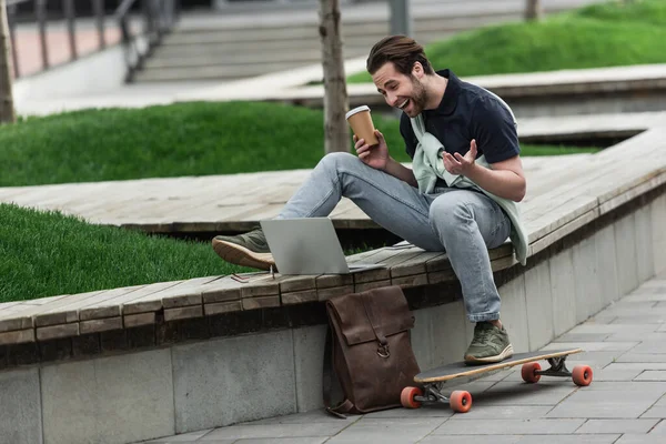 Cheerful man in polo shirt and sweatshirt holding coffee to go and laughing while sitting near longboard and gadgets — Stock Photo