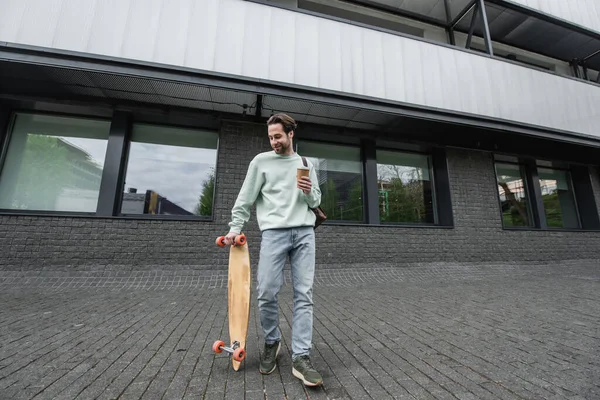 In voller Länge glücklicher Mann im Sweatshirt mit Pappbecher und Longboard draußen — Stockfoto
