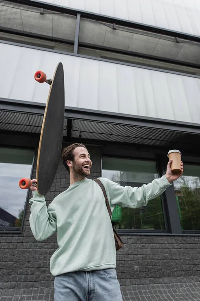 Allegro uomo in felpa tenendo tazza di carta e longboard al di fuori — Foto stock