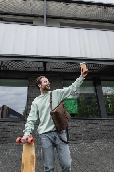 Smiling man in sweatshirt holding paper cup and longboard outside — Stock Photo