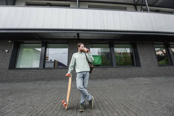 Pleine longueur de l'homme en sweat-shirt boire du café pour aller et tenant longboard à l'extérieur — Photo de stock