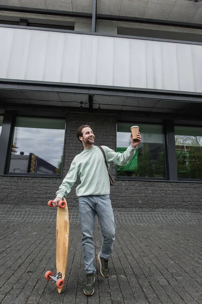 Comprimento total do homem alegre no sweatshirt segurando copo de papel e longboard fora — Fotografia de Stock