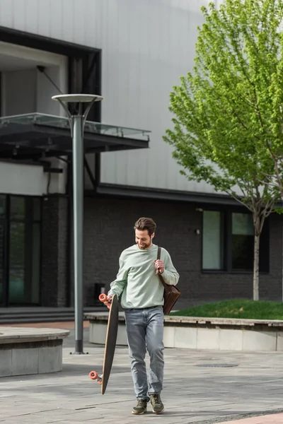 Full length of pleased bearded man in sweatshirt holding leather strap of backpack and longboard while walking outside — Stock Photo