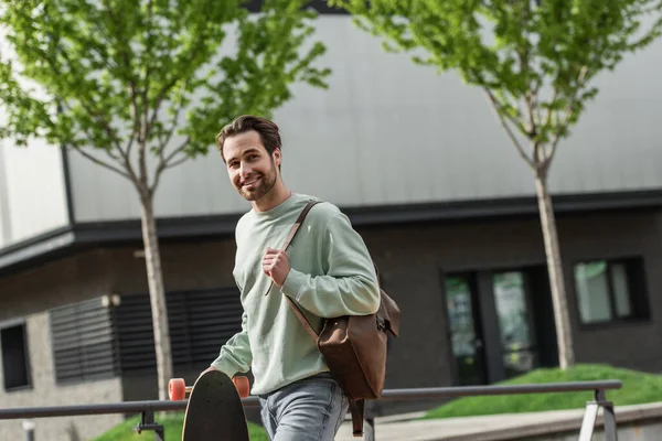 Glücklicher bärtiger Mann im Sweatshirt mit Lederriemen von Rucksack und Longboard draußen — Stockfoto