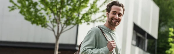 Усміхнений бородатий чоловік у сорочці тримає шкіряний ремінь рюкзака біля будівлі зовні, банер — стокове фото