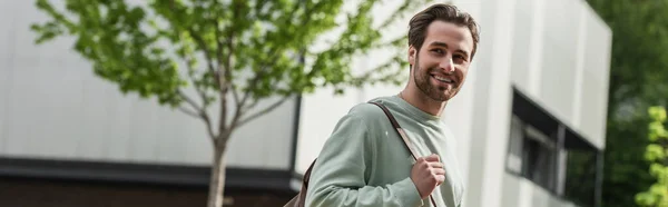 Hombre barbudo sonriente en sudadera con correa de cuero de la mochila cerca del edificio exterior, pancarta - foto de stock